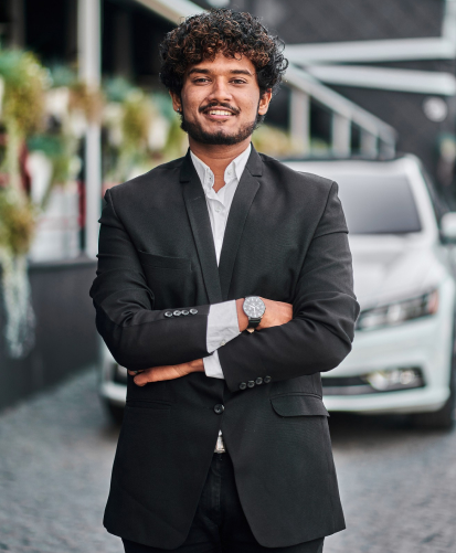 "Professional young Indian businessman in black suit smiling confidently with crossed arms against modern office background"