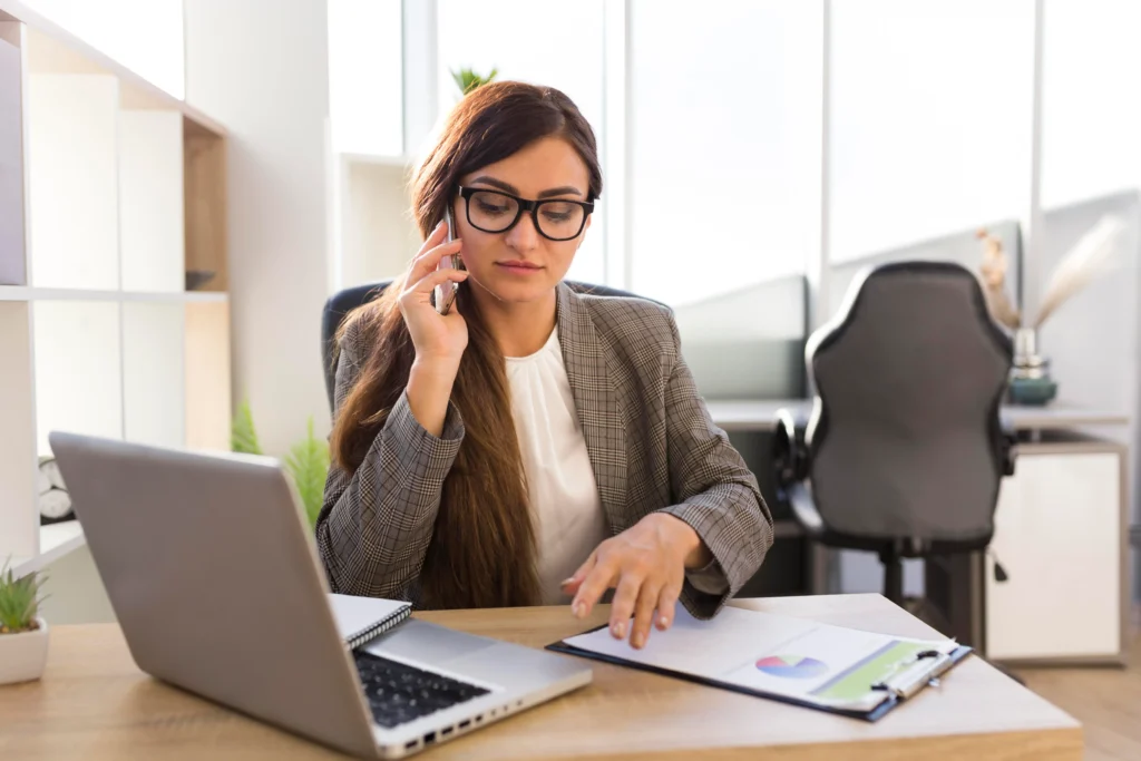 "Professional businesswoman in glasses taking phone call while reviewing financial reports at modern office desk"