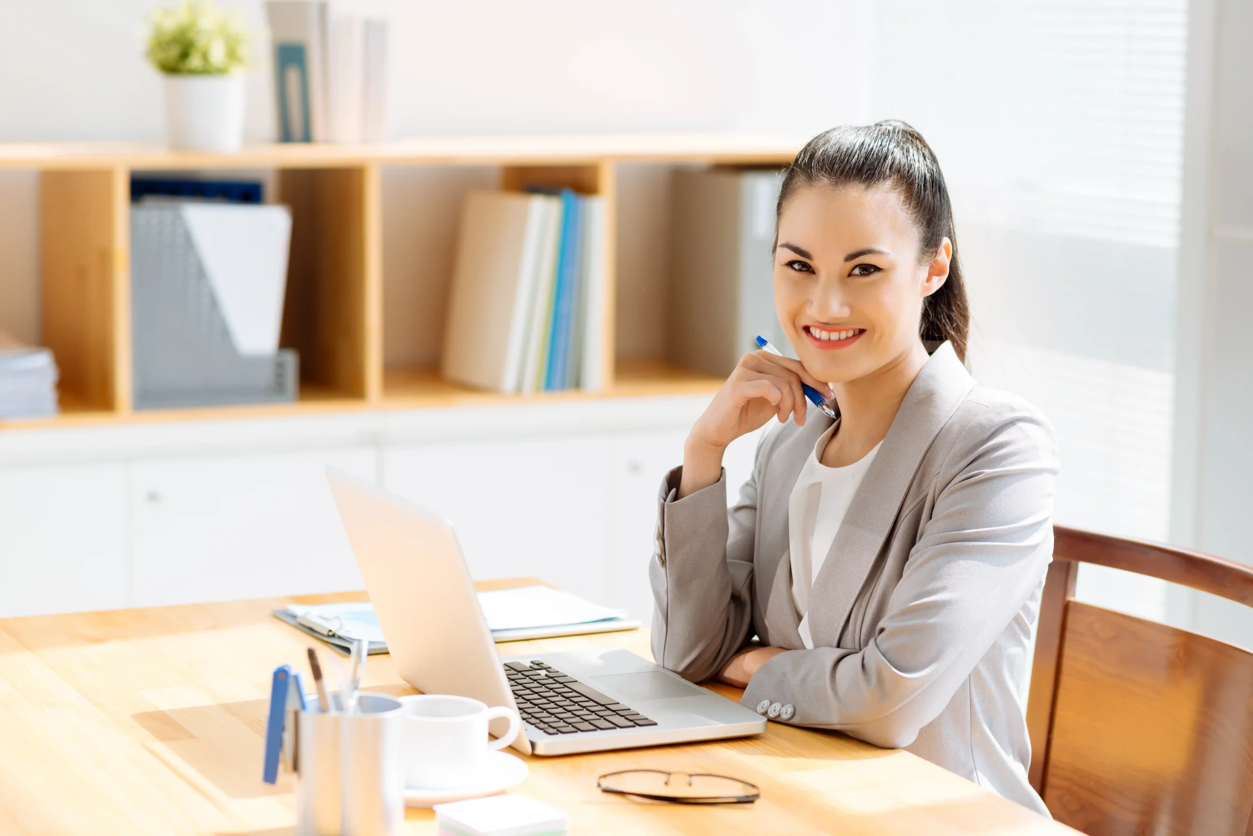 "Professional Asian businesswoman smiling at desk with laptop in modern office workspace"