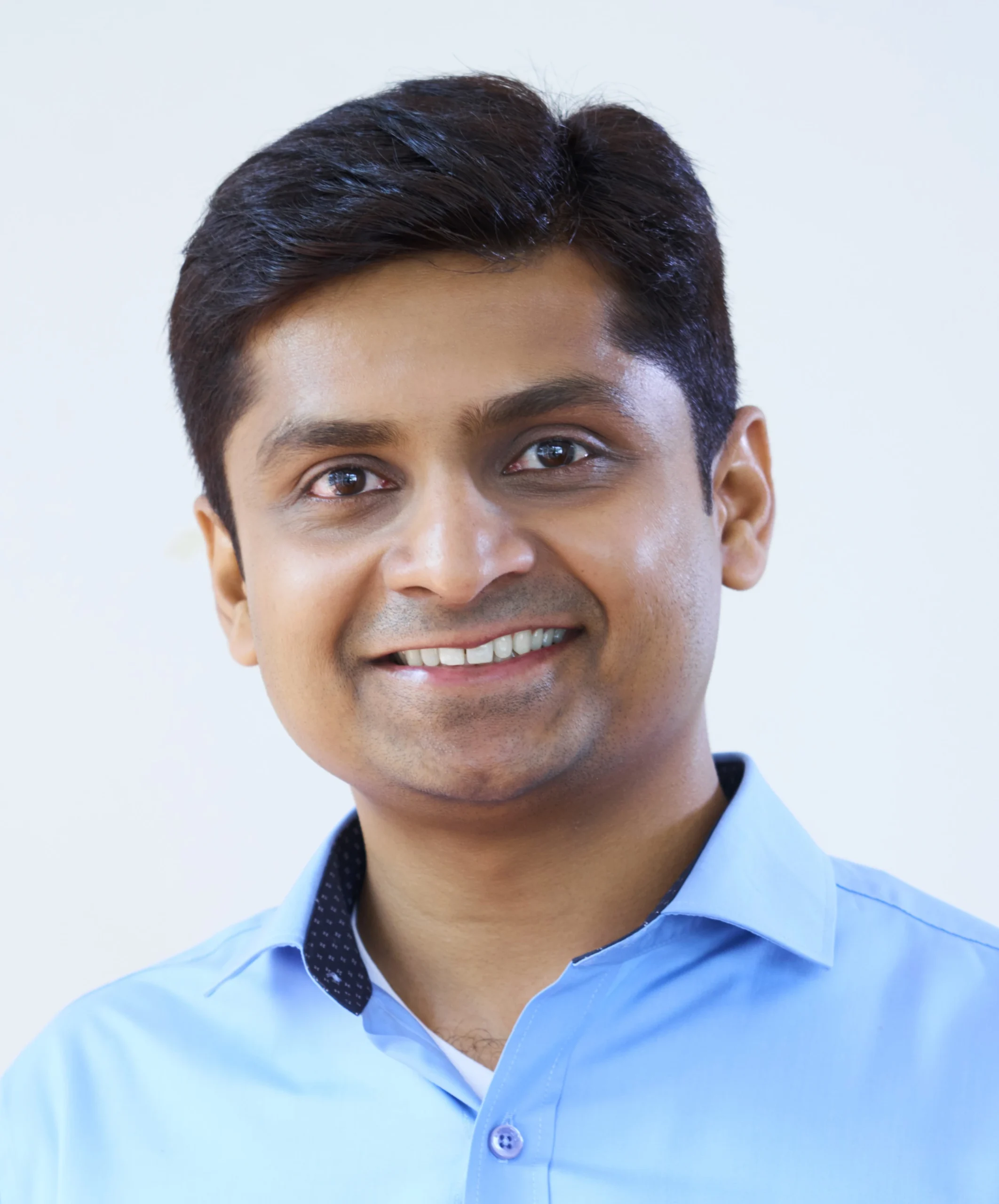 "Professional headshot of young Indian businessman wearing light blue collared shirt smiling against white background"